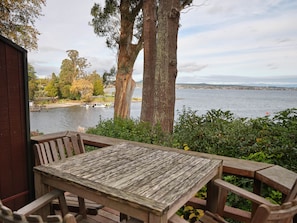A wooden deck with a table and benches overlooking a lake with boats, surrounded by trees and a distant cityscape.