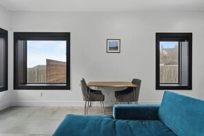A room with a wooden dining table, two chairs, a blue sofa, and two large windows with black frames.