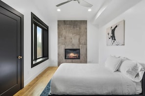 A bedroom with a bed, a fireplace, a ceiling fan, and a large window.