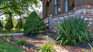A brick pathway leading to a building with a large window and a well-maintained garden with various plants.