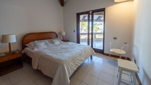 Ein Schlafzimmer mit einem hölzernen Bett, einem Nachttisch mit Lampe, einem Sessel und Blick ins Freie durch eine Glastür.