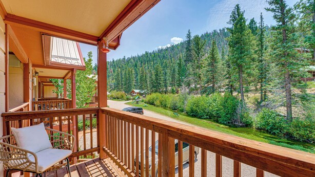 A wooden balcony with a wicker chair and a table, overlooking a forested area with a road and a car.