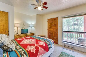 A bedroom with a bed, a ceiling fan, a TV on a wooden dresser, a door, and a balcony with a railing. - Bike, Hike & Ski: Creek-View Condo in Red River! (Red River)