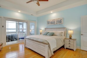 Une chambre à coucher avec un lit, des tables de chevet, un ventilateur de plafond et une vue sur l’extérieur, accessible par une porte-fenêtre coulissante.