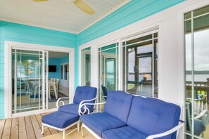 Un espace extérieur abrité, aux couleurs bleu et blanc, comprenant un canapé et des fauteuils bleus, une porte vitrée donnant sur un espace intérieur et une terrasse en bois.