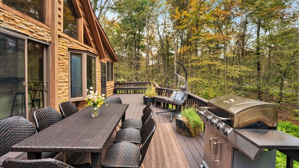 Une terrasse en bois avec un espace repas, un barbecue extérieur et de grandes fenêtres donnant sur une forêt.