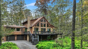 Une cabane en rondins à deux étages, dotée d’une cheminée en pierre, d’une terrasse et d’une allée en gravier, le tout entouré d’une végétation luxuriante composée de fougères et d’arbres.
