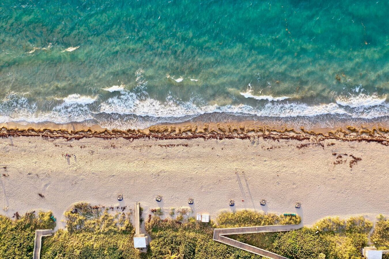 Vista aerea di una spiaggia con acque turchesi cristalline, una spiaggia sabbiosa e alcuni edifici, tra cui un sentiero e piccole costruzioni.