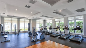 A spacious gym with multiple treadmills, elliptical machines, and weight stations. Large windows provide natural light and views of greenery outside.