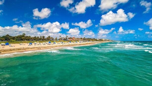 A beach with clear turquoise waters, a sandy shore, and a row of buildings along the coastline.