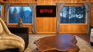 A cozy living room with a wooden TV wall, a large wooden coffee table, and a view of trees through the windows. - Rustic Log Cabin near National Park with Hot Tub & Mountain Views (Allenspark)