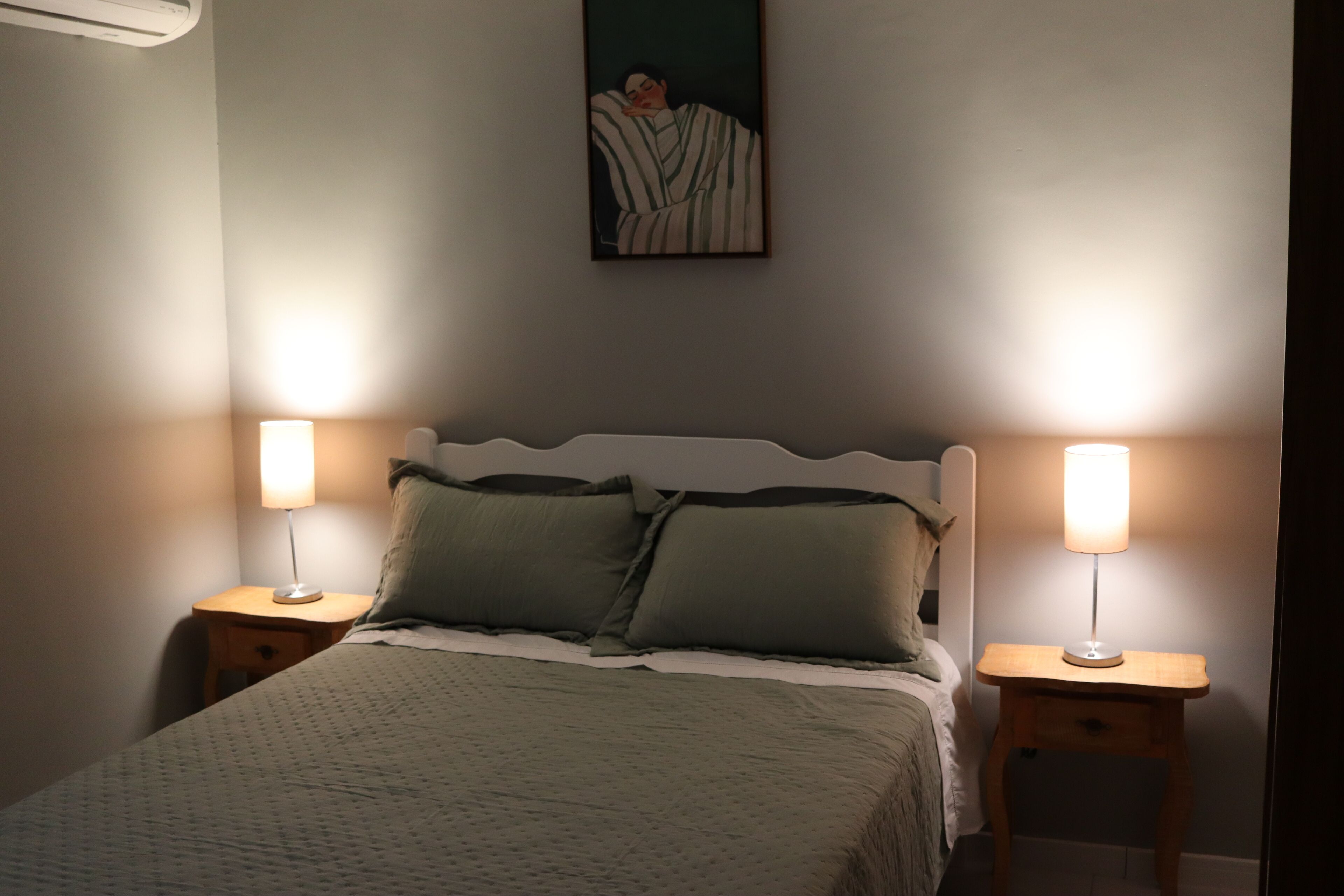 Un lit bien fait, avec deux tables de chevet, chacune surmontée d’une lampe, dans une pièce dont le mur est orné d’un seul tableau.