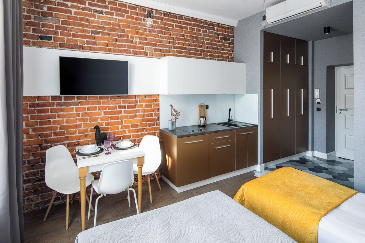 A modern hotel room with a brick wall, a flat-screen TV, a kitchenette, a dining area with a table and chairs, and a bed with a yellow blanket.