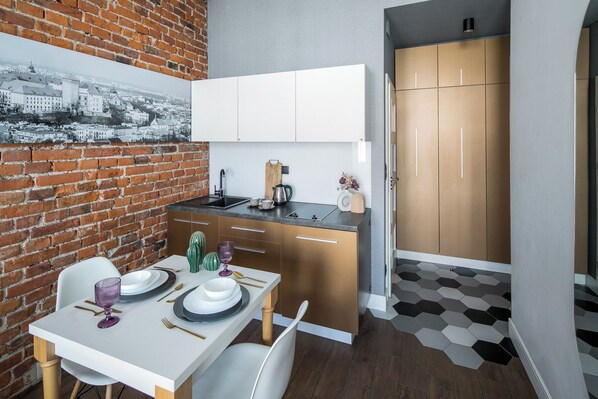 Une cuisine compacte avec un mur en briques, un coin repas avec une table blanche dressée pour deux personnes, et un couloir avec un sol à motifs géométriques.