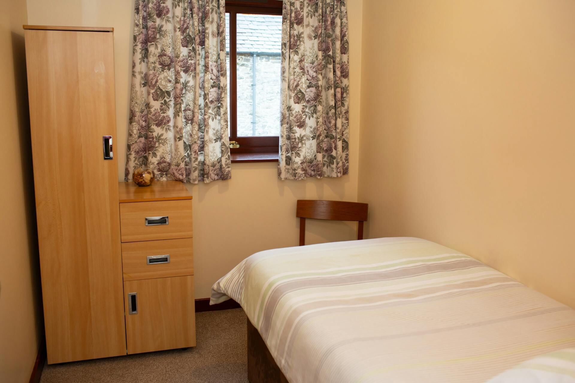 Un dormitorio con un armario de madera, una cama con colcha a rayas y cortinas florales en una ventana.