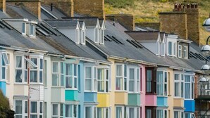 A row of colorful terraced houses with pitched roofs and chimneys, set against a green hillside.