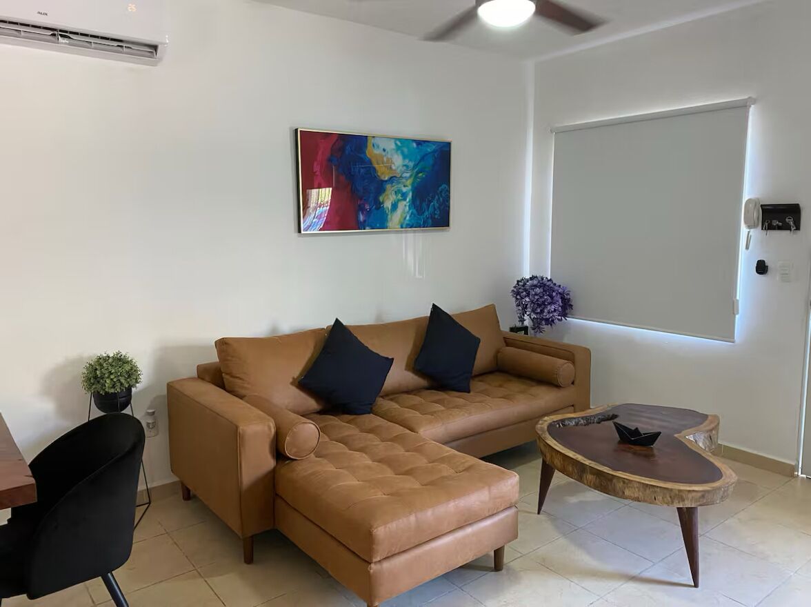 A living room with a brown leather sofa, a small round coffee table, a black chair, a colorful abstract painting, and a potted plant.