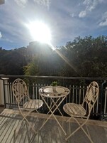 Un balcone con due sedie di metallo bianco e un tavolino rotondo, una pianta in vaso e vista su un edificio e alberi sotto un cielo soleggiato.