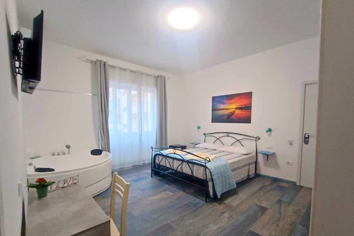 A bedroom with a bed, a bedside table, a chair, a window with curtains, a painting on the wall, and a bathroom with a sink and mirror.