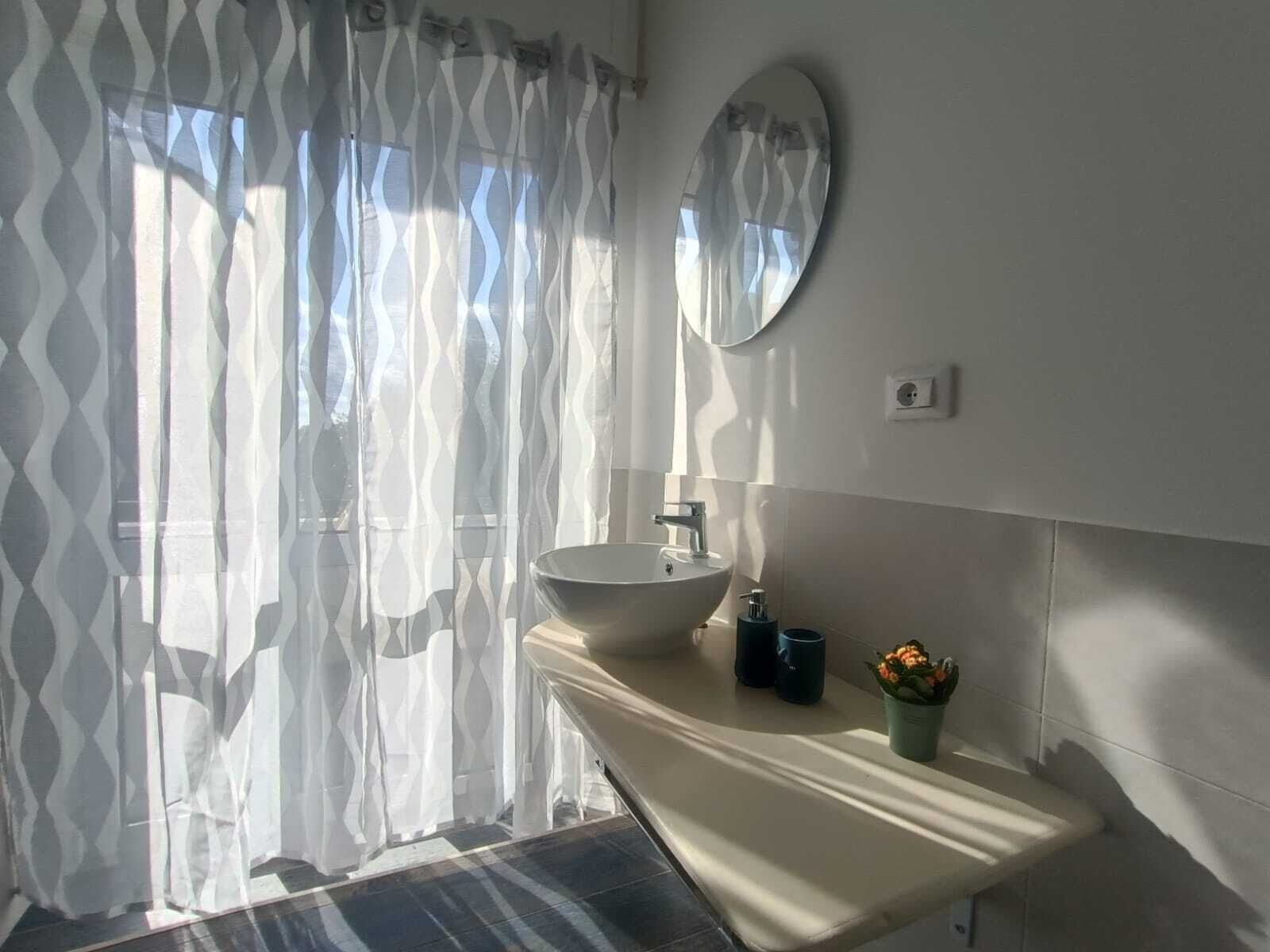 A bathroom with a white sink, a mirror, and a window with sheer curtains.