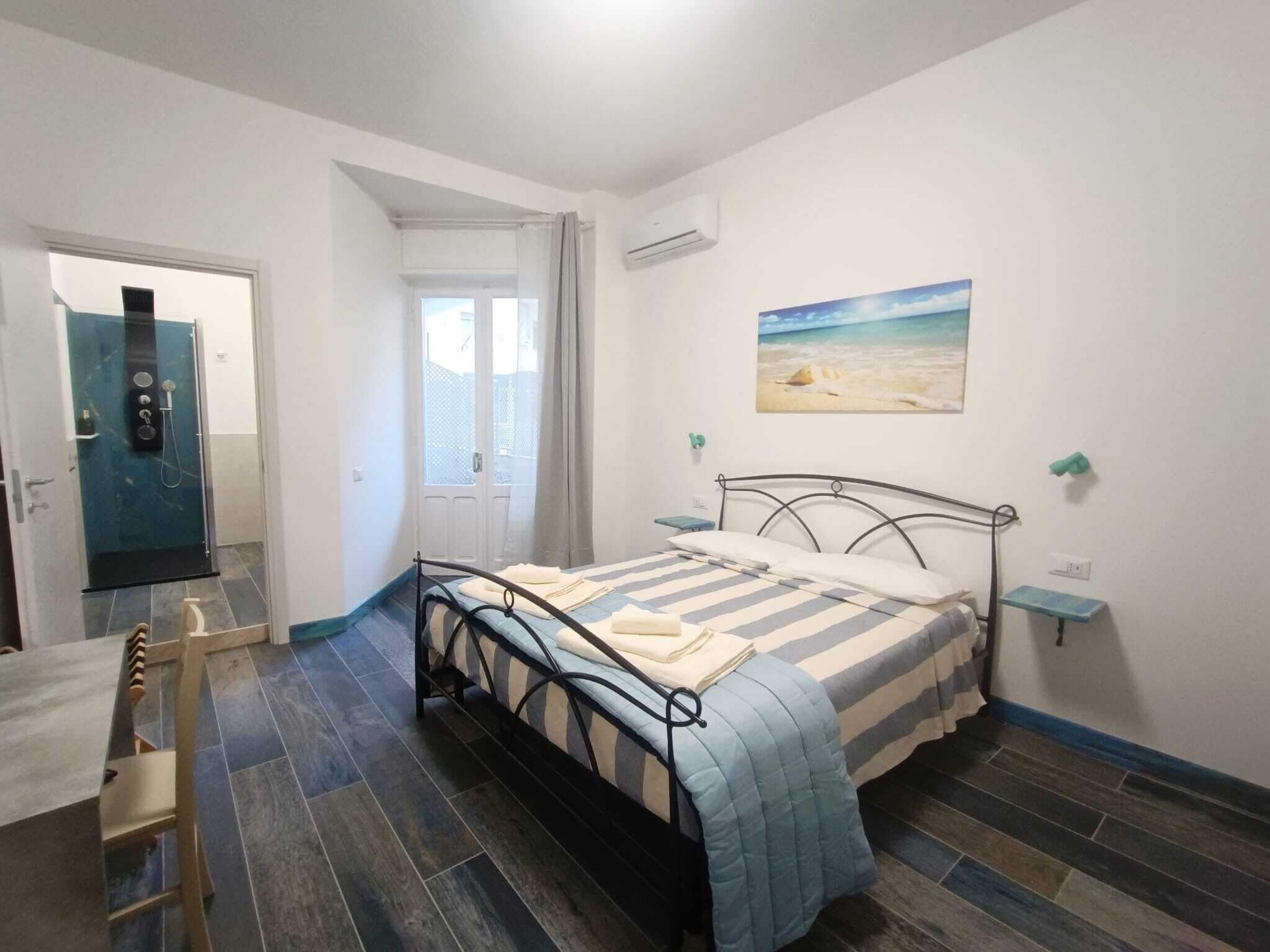 A bedroom with a bed, a window, a door, and a painting of a beach.