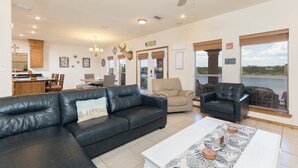 Ampla sala de estar com sofá de couro preto, mesa de centro branca e ventilador de teto. Há uma área de jantar com mesa e cadeiras de madeira, e uma cozinha com armários de madeira.