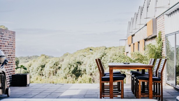 An outdoor patio with a table and chairs, a brick wall, and a view of a hilly landscape. - Alkina Lodge Milu Great Ocean Road (Wattle Hill)