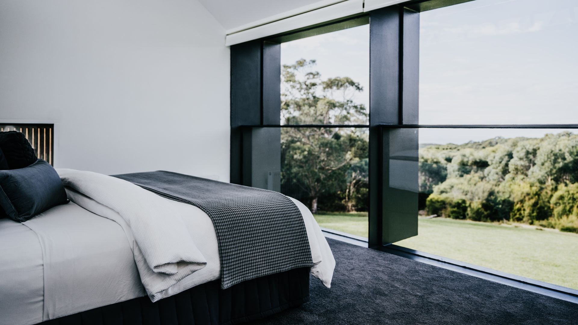 A modern bedroom with a large window, a bed with a black headboard, and a view of a green landscape.