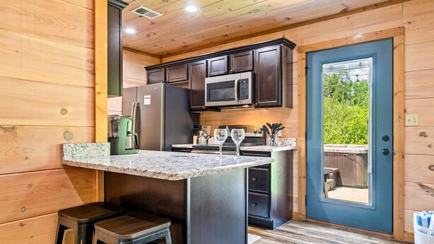 A compact kitchen with dark wood cabinets, a stainless steel refrigerator, a microwave, and a granite countertop. There are two stools and a door leading to an outdoor area. - Close to Dollywood-Hot Tub & Kids Closet (Pigeon Forge)