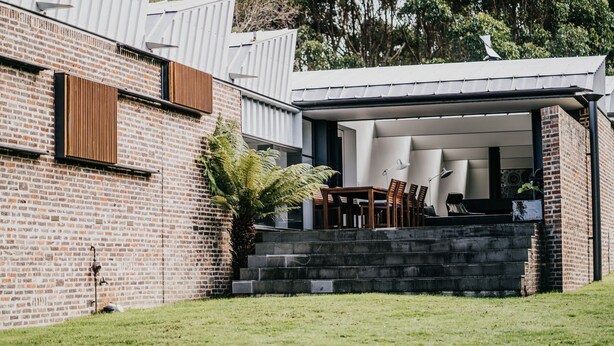 A modern house with a brick exterior, a covered outdoor seating area with a table and chairs, and a large fern plant. - Alkina Lodge Nuru (Wattle Hill)