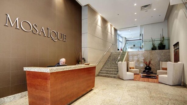 Un comptoir d’accueil avec une enseigne indiquant « MOSAIQUE », situé dans un hall moderne agrémenté d’un escalier, d’un espace salon et d’une rambarde en verre.
