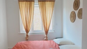 A bedroom with a bed, two windows with curtains, a radiator, and a small bedside table.