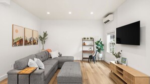 Ein modernes Wohnzimmer mit einem grauen Ecksofa, einem Holztisch, einem an der Wand montierten Fernseher und einem Bücherregal mit Pflanzen.