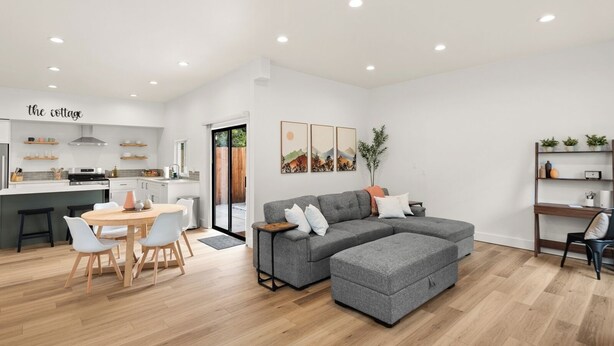Ein modernes Wohnzimmer mit einem grauen Ecksofa, einem Holzesstisch mit Stühlen und einer Küchenzeile mit weißen Schränken und einem Herd.