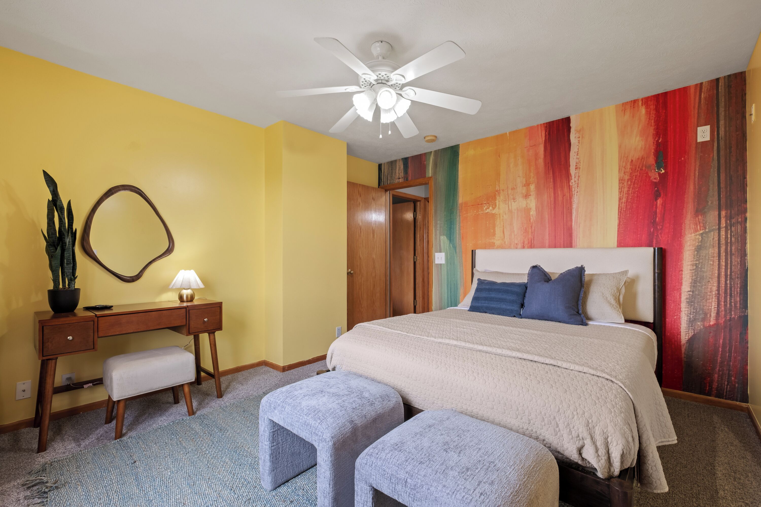 A bedroom with a bed, a desk, a chair, a ceiling fan, and a colorful wall mural.