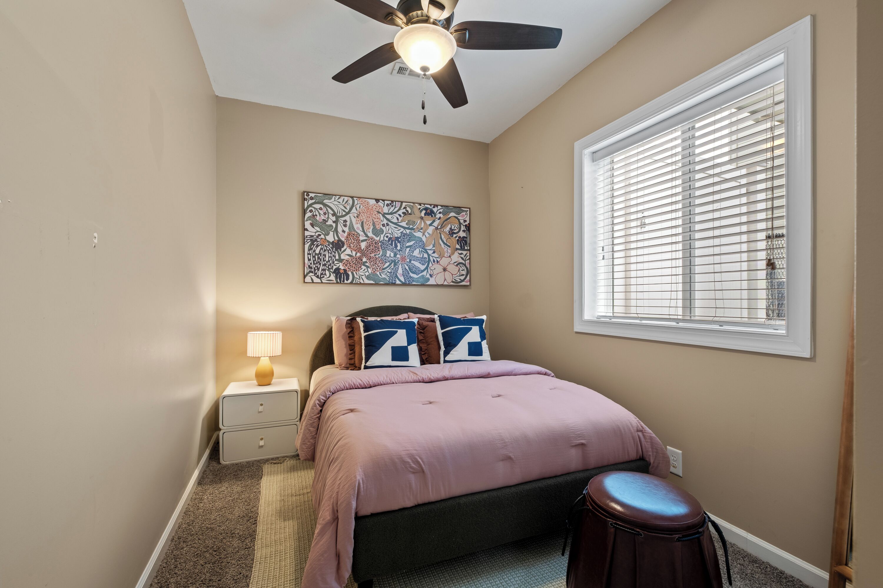 A bedroom with a bed, a nightstand, a ceiling fan, a window with blinds, and a wall-mounted artwork.