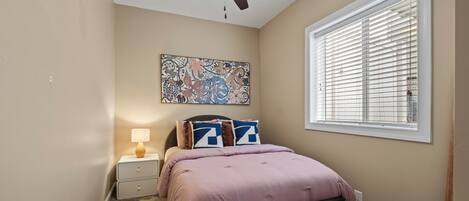 A bedroom with a bed, a nightstand, a ceiling fan, a window with blinds, and a wall-mounted artwork.