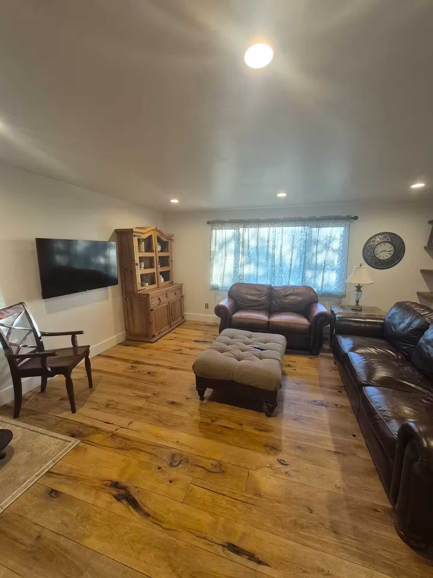 A living room with a wooden floor, a TV mounted on the wall, a leather sofa, a wooden cabinet, and a window with curtains.