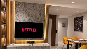 A modern living room with a large flat-screen TV mounted on a marble wall, a wooden entertainment unit, a dining area with yellow chairs, and a painting on the wall. - Beautiful 2-bedroom apartment in the heart of Gueliz (Marrakech)