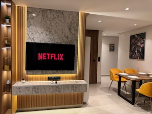 A modern living room with a large flat-screen TV mounted on a marble wall, a wooden entertainment unit, a dining area with yellow chairs, and a painting on the wall.