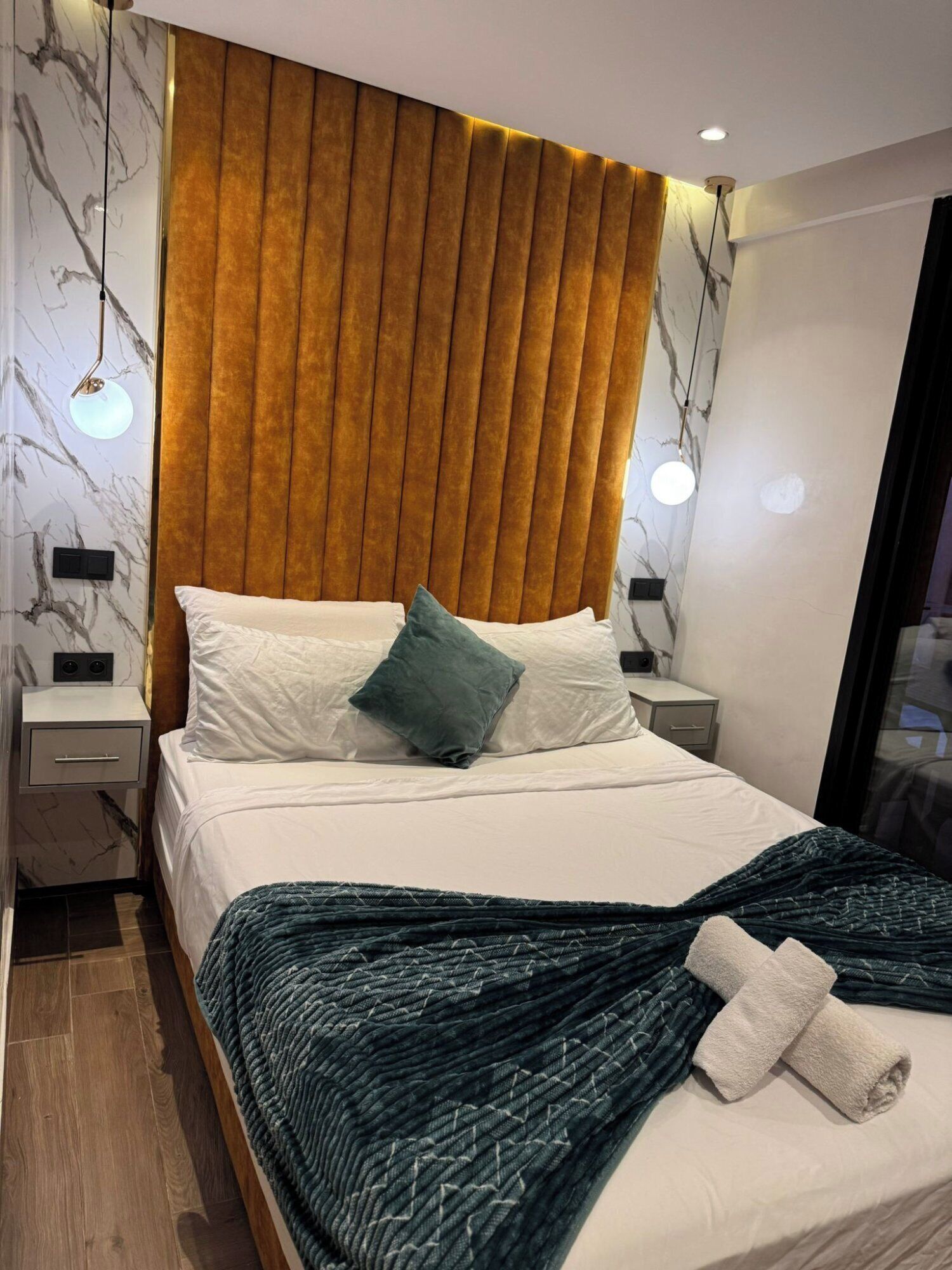 A modern hotel room with a large bed, a headboard with vertical slats, bedside lamps, and a marble-patterned wall.