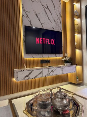 A modern living room with a large flat-screen TV displaying 'NETFLIX,' a marble-patterned fireplace, and a tray with glasses and a teapot.