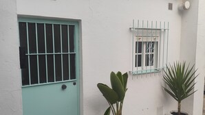 Un edificio blanco con una puerta verde azulada, una ventana con reja metálica y dos macetas con plantas.