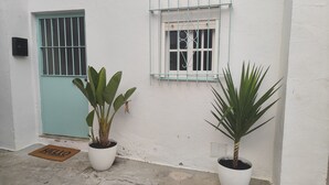 Un edificio blanco con una puerta azul y una ventana con celosía metálica. Hay dos plantas en macetas frente al edificio.