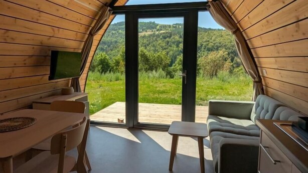 An interior with a wooden sloped ceiling, a dining area with a table and chairs, a sofa, and a kitchenette with cabinets.