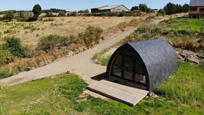 A modern, dome-shaped structure with a wooden deck, situated in a rural landscape with a dirt path leading to it.