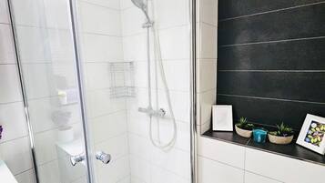 A modern bathroom with a glass-enclosed shower, white tiled walls, a black accent wall, and a shelf with small plants and a framed picture.