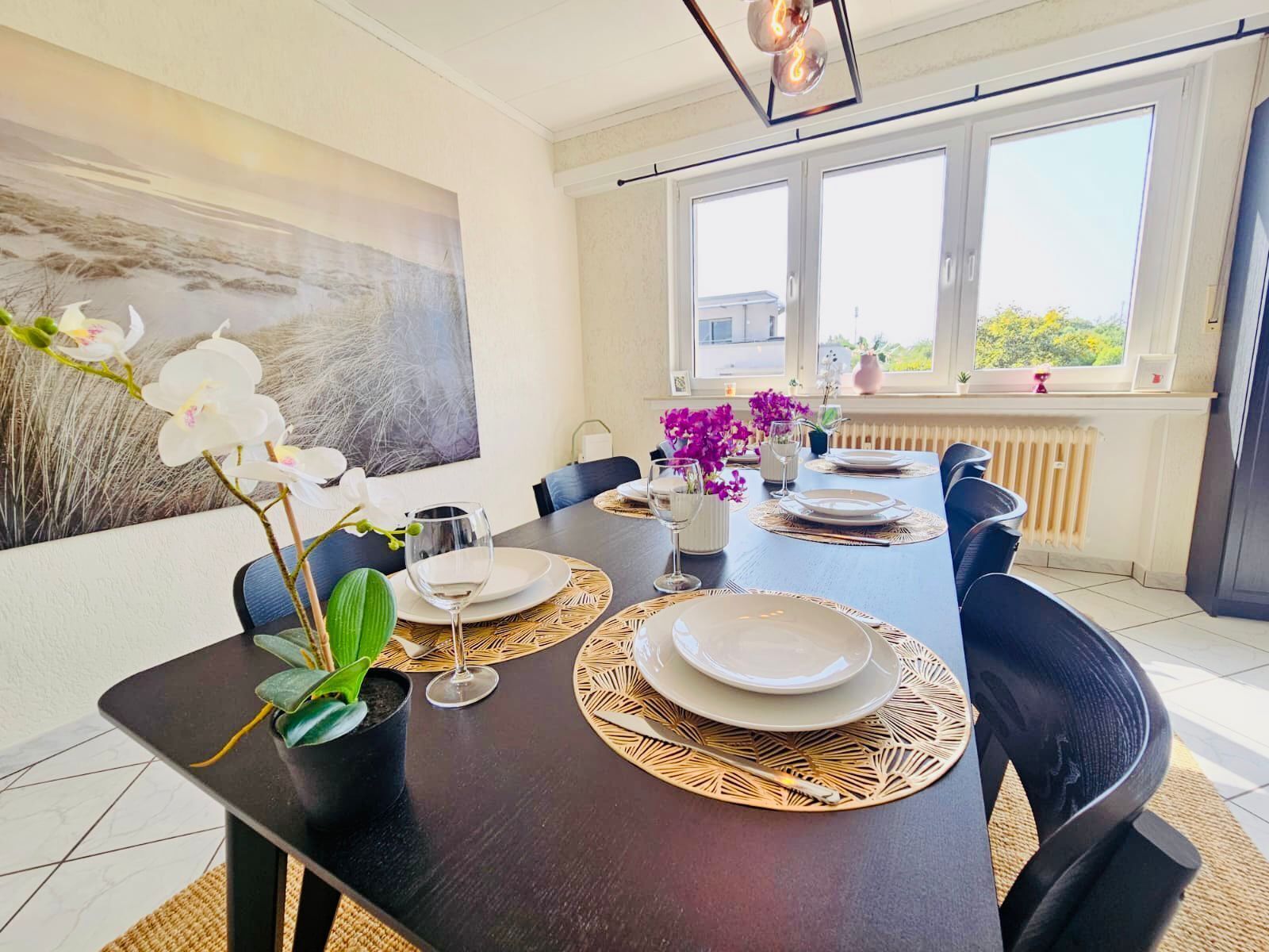 Un comedor con una mesa de madera oscura dispuesta para cuatro personas, rodeada de sillas. Hay dos ventanas que proporcionan luz natural, y un radiador debajo.