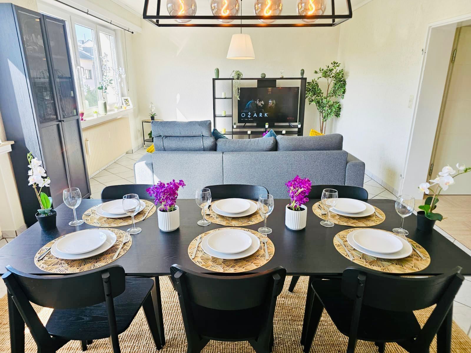 A dining area with a black table set for four, surrounded by black chairs. There is a grey sofa in the background, a television on a stand, and a potted plant on the right.