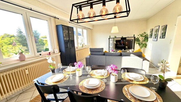 A dining area with a table set for four, a black sofa, a TV, and a view of greenery outside. - Chic Urban Stay 2BR with parking (Luxembourg City)